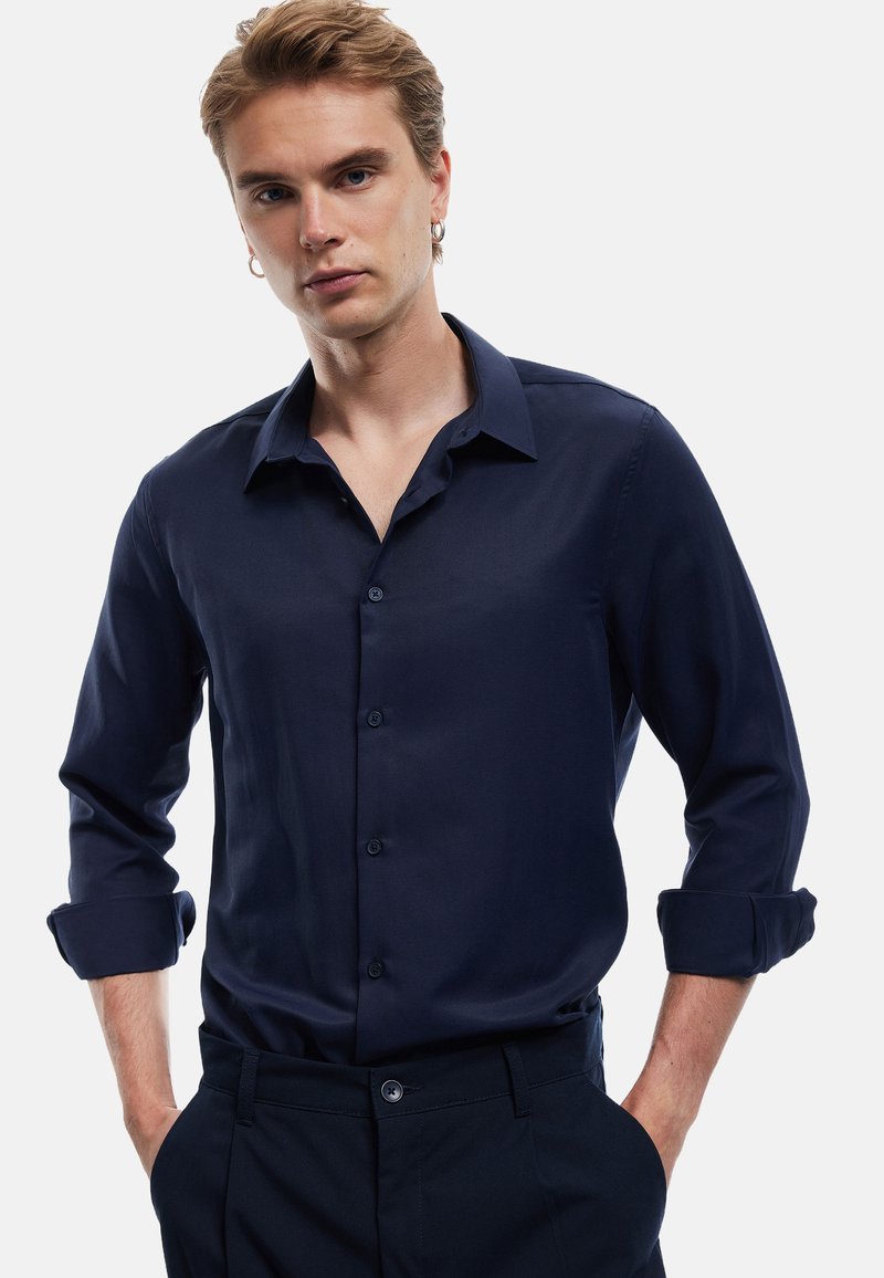 Camicia bottonata blu navy con maniche lunghe arrotolate ai polsini, realizzata in tessuto morbido, con colletto classico e una leggera lucentezza.
