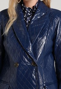 Blazer trapuntato blu navy realizzato in materiale lucido, con una chiusura doppiopetto e bottoni tondi, abbinato a una sciarpa a pois sottostante.