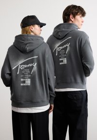 Δύο άτομα που φορούν γκρι φούτερ με κουκούλα με τη λέξη «Tommy» και ονόματα πόλεων τυπωμένα στην πλάτη, στέκονται δίπλα-δίπλα με την πλάτη τους προς την κάμερα.