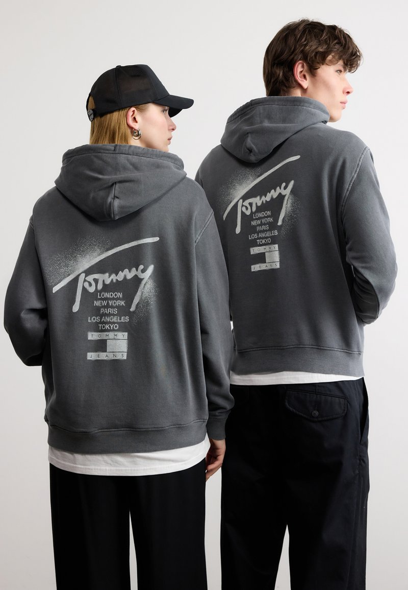 Δύο άτομα που φορούν γκρι φούτερ με κουκούλα με τη λέξη «Tommy» και ονόματα πόλεων τυπωμένα στην πλάτη, στέκονται δίπλα-δίπλα με την πλάτη τους προς την κάμερα.