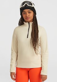 Beige fleece-pusero, jossa on puolilukko ilmanvaihtoa varten. Musta vetoketju yksityiskohtia, jotka sisältävät logon, yhdistettynä oransseihin kapea malli housuihin ja mustaan pipoon.