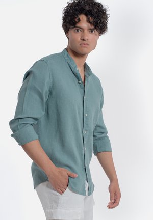 Giovane uomo con capelli ricci che indossa una camicia verde acqua con bottoni e pantaloni bianchi, con una mano in tasca, in piedi davanti a uno sfondo bianco.