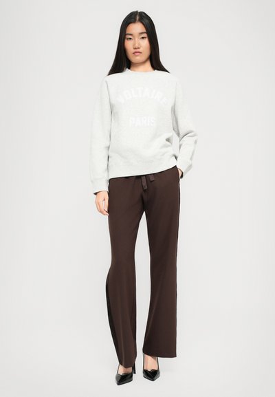 Jeune femme aux longs cheveux noirs portant un sweat-shirt Voltaire Paris gris clair, un pantalon marron foncé et des escarpins noirs à talons aiguilles, se tenant droite.