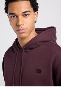 Joven con cabello corto y rizado que lleva una sudadera con capucha de color granate oscuro con cordones y un pequeño bordado de un logo en forma de diamante en el pecho.