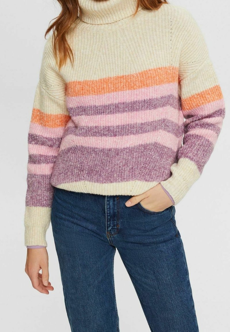 Femme portant un pull à col roulé crème avec des rayures horizontales orange, rose et violette, associé à un jean bleu, main dans la poche.