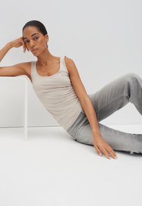 Frau lehnt sich auf dem Boden zurück, ein Ellbogen auf weißem Tisch, trägt ein beiges Tanktop und hellgraue Jeans vor einfachem weißem Hintergrund.