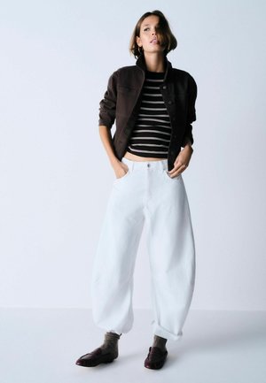 Nő áll, kezét zsebében tartja, fekete rövidített kabátot, fekete-fehér csíkos felsőt, széles fehér nadrágot, szürke zoknit és sötét loafert visel.