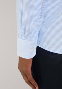 Camicia a maniche lunghe blu e bianca a righe con una texture liscia; presenta un polsino con bottoni e un rivestimento azzurro in contrasto.