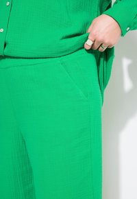 Ensemble en tissu vert texturé comprenant une chemise et un pantalon ; la chemise a des boutons et une coupe ample ; le pantalon présente des poches latérales et une silhouette décontractée.