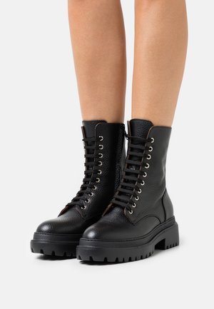 Botas de tobillo de cuero negro con un acabado texturizado, que presentan un diseño con cordones, ojales de metal y una suela de goma gruesa con un patrón de relieve.