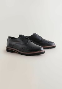 Čierne kožené brogues s ozdobnými perforáciami, šnurovacie zapínanie a podrážka s hnedým lemovaním na svetlo béžovom pozadí.