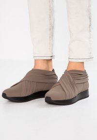 Scarpe in tessuto elastico marrone con design plissettato, punta arrotondata e suola in gomma nera, abbinate a jeans chiari con risvolto.