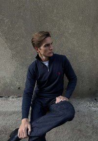 Fiatal férfi tengerészkék pulóverben és sötét farmernadrágban, egy texturált szürke falnak támaszkodva, oldalra nézve, egyik lábát a másik keresztezi.