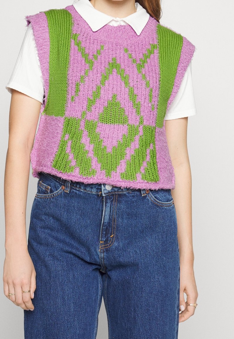 Pletená vesta v purpurové a zelené s geometrickými vzory, volného střihu a bez rukávů, nošená přes bílou košili s límečkem.