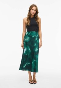 Haut sans manches noir associé à une jupe maxi en satin vert présentant un motif abstrait fluide. Sandales avec des lanières fines et des accents dorés.