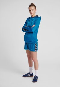 Turkosidrotts hooded sweatshirt med grå huva och svarta detaljer, ihop med matchande shorts med svarta chevronmönster. Blå sportskor.