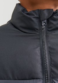 Črni puhovka z visokim ovratnikom, brezšivnim zapiranjem na zadrgo in mat teksturo tkanine. Oznaka z logotipom je vidna blizu zadrge.