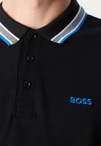 Polo noir avec trois boutons, col à rayures bleu et blanc, et logo "BOSS" brodé sur la poitrine.