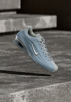 Chaussure de sport Nike bleu clair et grise avec amorti à air visible, flottant au-dessus d'une surface en béton avec des marches en arrière-plan.