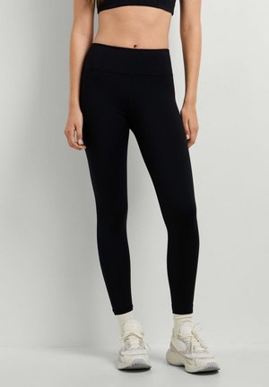 Vrouw draagt zwarte legging met hoge taille, witte sokken en witte sneakers, staand tegen een effen lichte achtergrond.