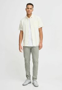 Korte mouwen, lichtgeel knopenhemd met een borstzak, gecombineerd met grijze broek en grijze sportschoenen, staande voor een effen achtergrond.
