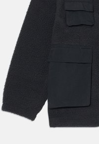 Svart fleecejakke med en strukturert overflate, som har en stor lappelomme og en glatt stofflomme, begge på nedre venstre side.