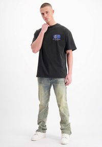 Zwarte katoenen t-shirt met een logo, losse fit, korte mouwen, gecombineerd met lichtblauwe distressed jeans en witte sneakers. Achtergrond is effen wit.