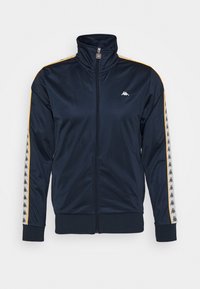 Marinblå zip-up träningsjacka tillverkad av slät tyg; har gula sidostrekar och en logotyp på bröstet, med elastiska ärmslut och fåll.