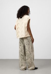 Kremowy, dzianinowy top bez rękawów o falistej teksturze, z czarną metką, zestawiony z luźnymi, marmurkowymi zielonymi spodniami z bocznymi kieszeniami.