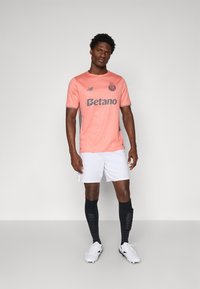 Camiseta de fútbol rosa con detalles en gris, pantalones cortos blancos, medias negras y botas blancas. Presenta el logo del patrocinador y el emblema del equipo en el pecho.