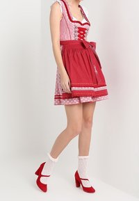 Robe dirndl rouge avec un corsage ajusté, un chemisier blanc contrastant, une jupe à motifs et un détail nœud. Associée à des chaussettes blanches en dentelle et des escarpins rouges.
