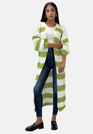 Długi kardigan w białe i zielone paski, dzianinowy materiał, otwarty z przodu, z opuszczonymi ramionami i ściągaczami na mankietach, zestawiony z dopasowanymi jeansami i mokasynami.