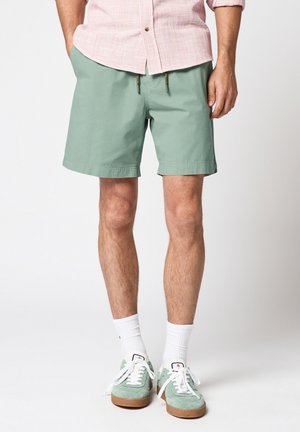 Uomo che indossa pantaloncini verde menta, scarpe da ginnastica bianche con suole di gomma, calzini bianchi e una camicia rosa con bottoni e maniche arrotolate.