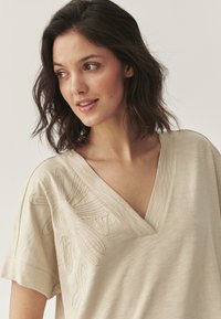 T-shirt beige en V en tissu doux, avec broderie florale sur le côté gauche. Manches courtes, coupe décontractée.