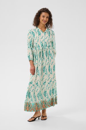 Femme portant une longue robe crème à motifs floraux verts et un ourlet décoratif orange, chaussée de sandales à talons noires, debout et souriante.