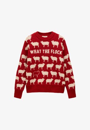 Sarkans adīts džemperis ar baltām aitu rakstiem un uzrakstu "KAS TAS PAR SŪNI" uz krūtīm, kurā ir viena melna aita starp baltajām aitu.