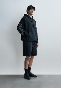 Jauns vīrietis valkā melnu kapuci ar baltām grafiskām piedurknēm, melnus šortus, melnu cepuri, augstas melnas zeķes un melnus sporta apavus, stāv pret vienkāršu sienu.