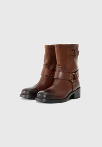 Bottines en cuir marron avec un bout arrondi, détail de sangle, accent de boucle et un talon noir solide. Texture lisse avec un éclat subtil.