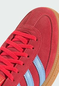 Detailný záber na červené semišové tenisky s jasne červenými šnúrkami, svetlomodrými cik-cak pruhmi a gumenou podrážkou.