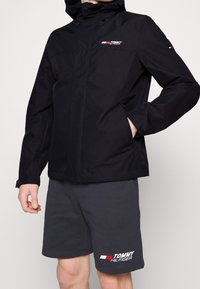 Sort vandtæt jakke med lynlås foran, justerbar hætte og logo på brystet; parret med grå shorts, der har et logo print.