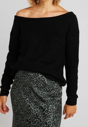 Kobieta ubrana w czarny sweter z odkrytymi ramionami i zieloną satynową spódnicę z czarnym wzorem w cętki lamparta.