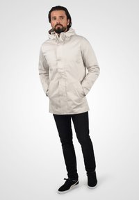 Lys beige vanntett jakke med hette, frontknapper, brystlomme og sidelommer, kombinert med svarte jeans og svarte joggesko.