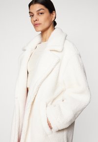 Cappotto in pelliccia sintetica bianca con ampio colletto, vestibilità oversize e design doppiopetto. Presenta tasche laterali e tessuto testurizzato.