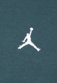 Ciemy zielony materiał z wytłoczonym białym logo Jumpman, przedstawiającym postać w powietrzu z piłką do koszykówki, tworzy teksturowany kontrast.