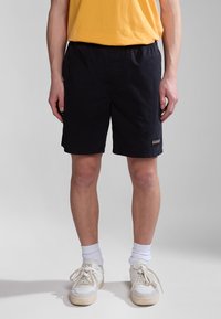 Pantalones cortos negros con una cintura elástica, hasta la rodilla, y un pequeño parche de logo en el costado. Llevados con zapatillas blancas y calcetines cortos.