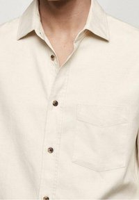 Camicia beige con bottoni, dotata di tasca anteriore, colletto aperto e bottoni marroni marmorizzati, indossata da una persona.