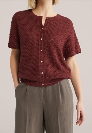 Femme portant un cardigan bordeaux texturé à manches courtes avec boutons et un pantalon gris ample taille haute, les mains dans les poches.