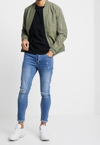 Olivgrüne Bomberjacke mit Reißverschluss, schwarzes Rundhals-T-Shirt, hellblaue, stark tragende Skinny-Jeans und weiße Sneaker.
