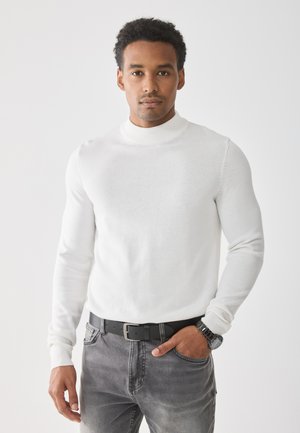 Maglione bianco a maniche lunghe con collo a imbuto in un morbido tessuto a maglia, abbinato a jeans grigi e una cintura nera. Orologio visibile al polso.