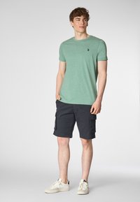 T-shirt verde a maniche corte con scollatura rotonda e un piccolo logo sul petto, abbinato a pantaloni cargo blu scuro e scarpe da ginnastica bianche.